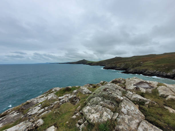 Cliffside view of the rugged Pembrokeshire coastline, West Wales. Overlooking a blue sea with white surf and green coastal grass and rocks on an overcast, summer's day. Viewed from the Wales coast path.