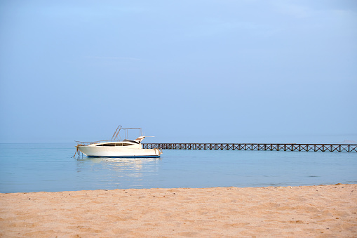 White motor boat floats in ocean water at long pier bridge under bright blue sky, view from sandy beach. Summer traveling concept.