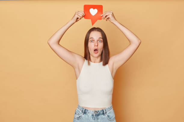 istock Blogging communication. Heart icon click. Online community interaction. Surprised excited brown haired young woman wearing casual clothing holding red like icon standing isolated over beige background 1676495570