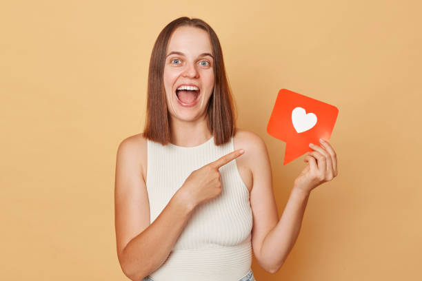 istock Vlog content. Love button. Web icon. Like notification. Smiling happy joyful brown haired young woman wearing casual clothing holding red like icon standing isolated over beige background 1676495574