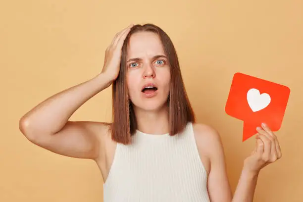 Photo of Heart communication. Like notification. Blogger recommendation. Shocked brown haired young woman holding red like icon standing isolated over beige background keeps hand on head looking with big eyes