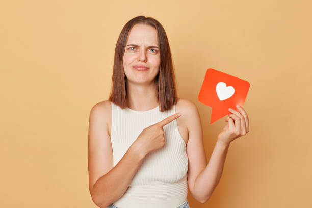 istock Like notification. Media networking. Comment counter. Confused brown haired young woman wearing casual clothing holding red like icon standing isolated over beige background 1676496105