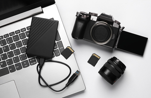 Laptop, external hard drive, modern digital camera with flip screen, two sd memory cards and lens on gray background. Photographer's equipment. Top view. Flat lay