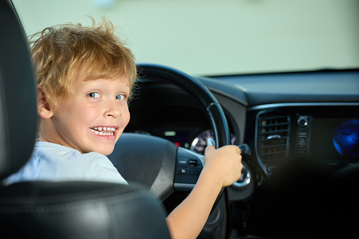 Funny boy turns the wheel in his father's car like a real driver. Child plays merrily with the steering wheel in the car. Five-year-old boy sits behind the wheel and pretends to drive a car