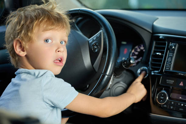 Blue-eyed boy is sitting in the driver's seat inside the car, pressed the ignition button with his finger. Boy looks back in fright while pressing the start button