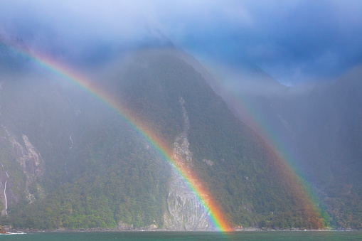 Photograph of a colourful and vibrant rainbow over the water in front of mountains in Milford Sound in Fiordland National Park on the South Island of New Zealand