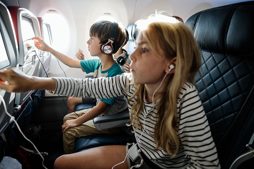 A Caucasian family with multiple children on vacation depart from the airport on a plane.  The children learn how to use the in flight entertainment to watch a movie.