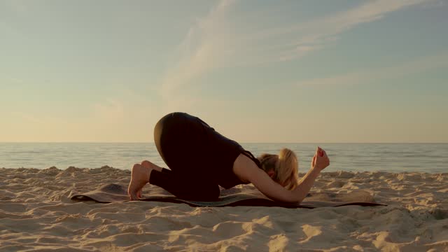 Middle years woman practicing asana yoga pose and sport training on fitness mat at sunset beach