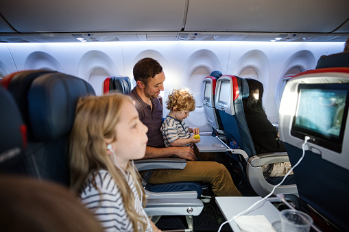 A Caucasian family with multiple children on vacation depart from the airport on a plane.  The dad carries his little boy on his lap for the flight.