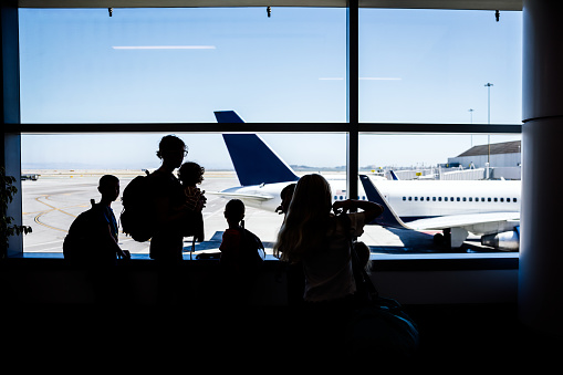 A  Caucasian family with multiple children traveling together at an airport, waiting to board their plane.  They look out a window at their plane, silhouetted by the light.