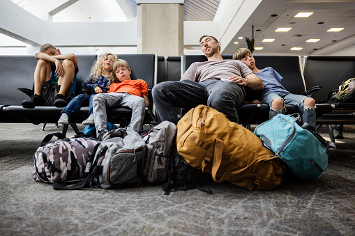 A  Caucasian family with multiple children traveling together at an airport, waiting to board their plane.  They sleep in a waiting area, surrounded by their luggage, bags, and carry on items.