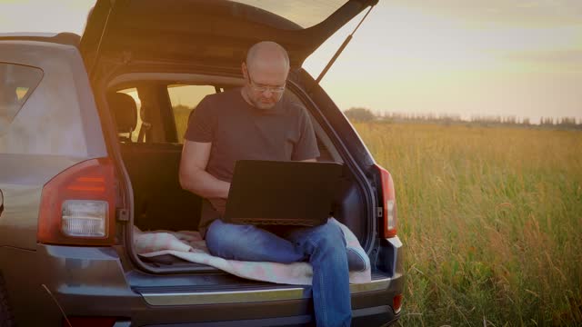Thoughtful worried man working remotely online typing internet communication technology sitting in car trunk while traveling
