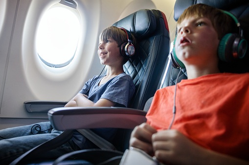 A Caucasian family with multiple children on vacation depart from the airport on a plane.  Two siblings ride next to each other, both wearing headphones for music or a movie.