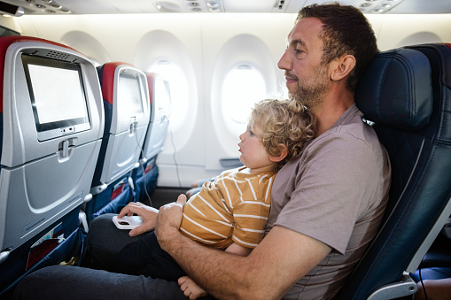 A Caucasian family with multiple children on vacation depart from the airport on a plane.  The dad carries his little boy on his lap for the flight.