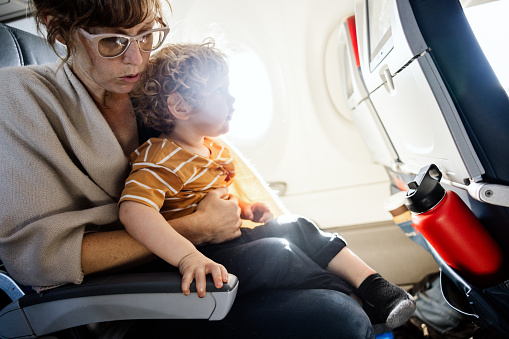 A Caucasian mother sits on a plane having just departed the airport, holding her toddler aged son on her lap.
