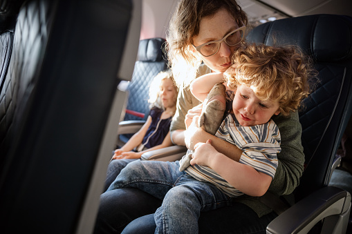 A Caucasian family with multiple children on vacation depart from the airport on a plane.  The mother holds a crying child experiencing ear pain on the flight.