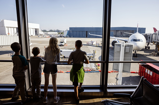 A Caucasian family with multiple children traveling together at an airport, waiting to board their plane.  The siblings look out the window at the boarding airplanes.