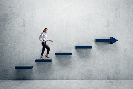 Side view of young woman climbing blue arrow stairs to success on concrete wall background in interior. Financial growth, career development and forward concept