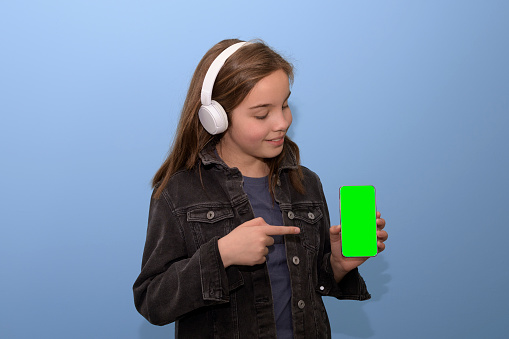 Green screen phone in the girl's hands. Portrait, blue background, teen girl in white bluetooth headphones