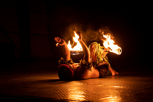 A Hawaiian male dancer holds a torch with his leg and claps his hands.