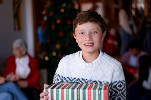 Portrait of beautiful 10 year old boy holding a Christmas present while facing the camera smiling - Family enjoying the Christmas celebration at background