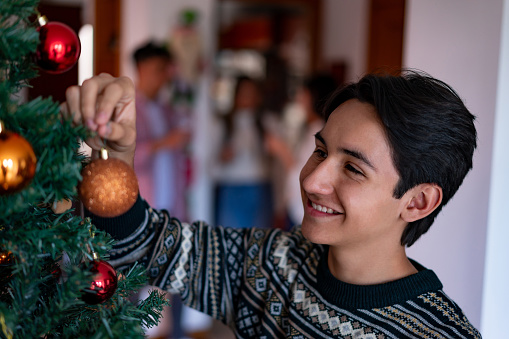 Handsome male teenager decorating the Christmas tree at home while looking very happy and smiling - Family enjoying the Christmas celebration at background
