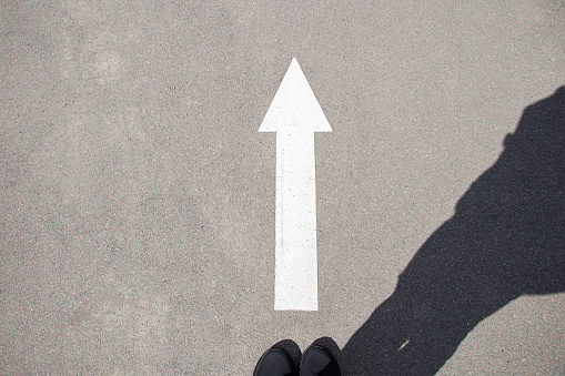 A woman in sneakers stands near a white arrow with a forward direction on the asphalt and a shadow of a woman, a road with a forward arrow
