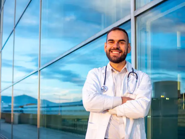 Male Doctor standing outside the hospital building with folded arms while looking at camera