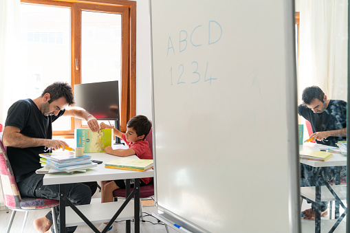 Father binding his son's books at his desk. Father and son are preparing for school together. There is a whiteboard in the foreground. It was shot with a full frame camera in daylight in the study room.