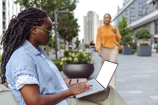 A young African American woman sitting on a bench and typing on her laptop, with another young African American woman walking in the background.