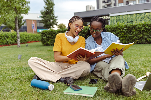 Two young African American female college students are talking and reading books together, while relaxing on a grass area.