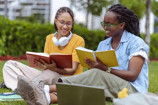 Two young African American female college students are sitting on a grass area and reading books together.