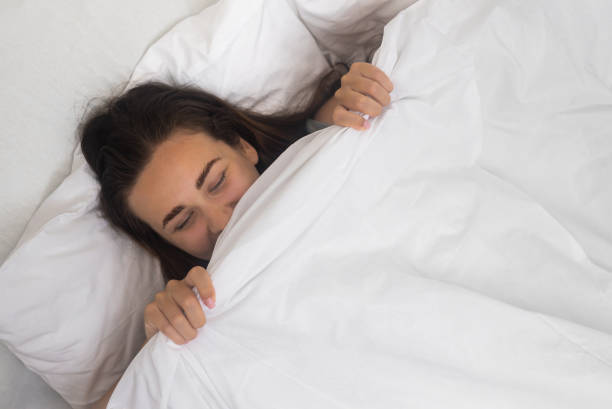 istock Young female laughs hiding face under blanket preparing for sleep in comfortable bed in evening 1676996853