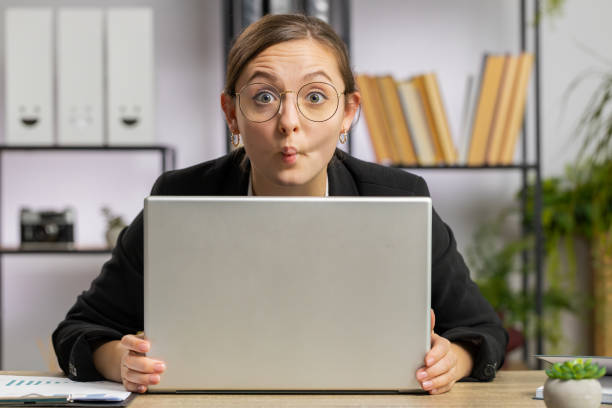 ラップトップコンピュータの後ろに隠れて、無礼な浮気をする面白い愚かな顔をするオフィスの実業家 - saleswoman glasses sales occupation bizarre ストックフォトと画像