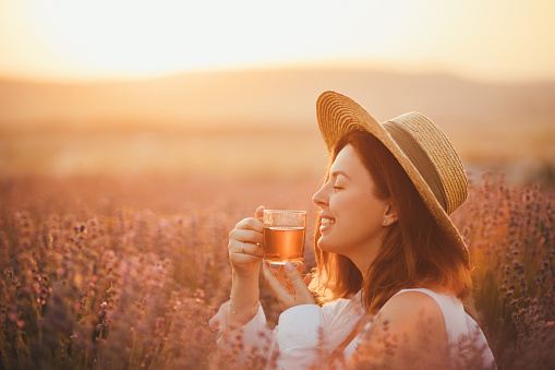 Young happy woman drinking healthy herbal tea, sitting in a beautiful lavender field at sunset.