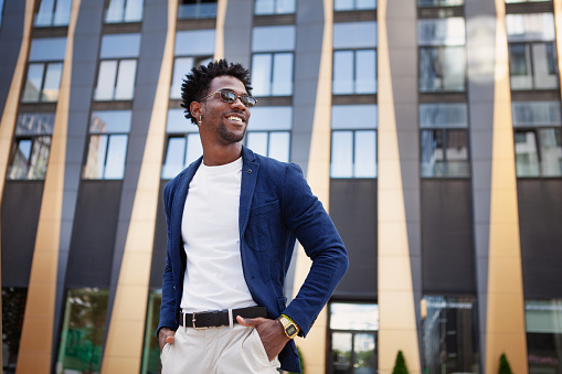 Confident african American male in stylish clothes stands in city next to modern building, looking to side with smile. Happy black man with hands in pockets.