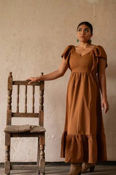 A Mexican woman graces an aged chair beneath delicate archways. Bathed in a play of light and shadow, her traditional attire and contemplative gaze