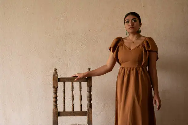 A Mexican woman graces an aged chair beneath delicate archways. Bathed in a play of light and shadow, her traditional attire and contemplative gaze