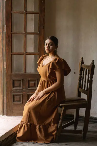 A Mexican woman graces an aged chair beneath delicate archways. Bathed in a play of light and shadow, her traditional attire and contemplative gaze