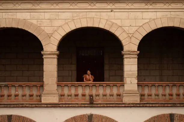 A Mexican woman graces an aged chair beneath delicate archways. Bathed in a play of light and shadow, her traditional attire and contemplative gaze