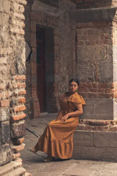 A Mexican woman graces an aged chair beneath delicate archways. Bathed in a play of light and shadow, her traditional attire and contemplative gaze