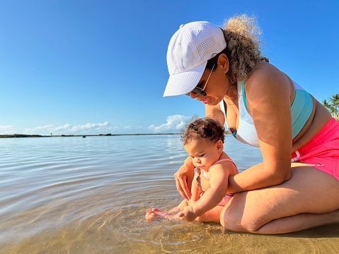 Baby girl and mother play with sand while in the water on a beach with coconut trees