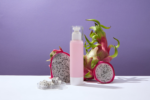 Fresh dragon fruit and dragon fruit slices decorated with a pink pump bottle on a purple background. Mockup product display for advertising. Vitamin C in dragon fruit helps skin stay youthful