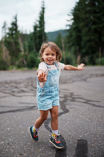 Happy and healthy preschool age Eurasian girl playfully holds out her arms like wings and runs through a parking lot before hiking with her family during a family road trip.