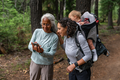 Active senior woman of Pacific Islander descent takes a break from hiking in a forest to show her adult daughter and one year old grandson a photo she took using her smart phone.