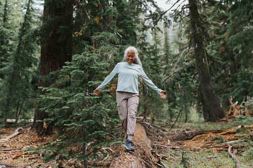 Active and healthy senior woman of Pacific Islander descent smiles while walking along the tree trunk of a fallen evergreen tree during a hike in the Pacific Northwest.