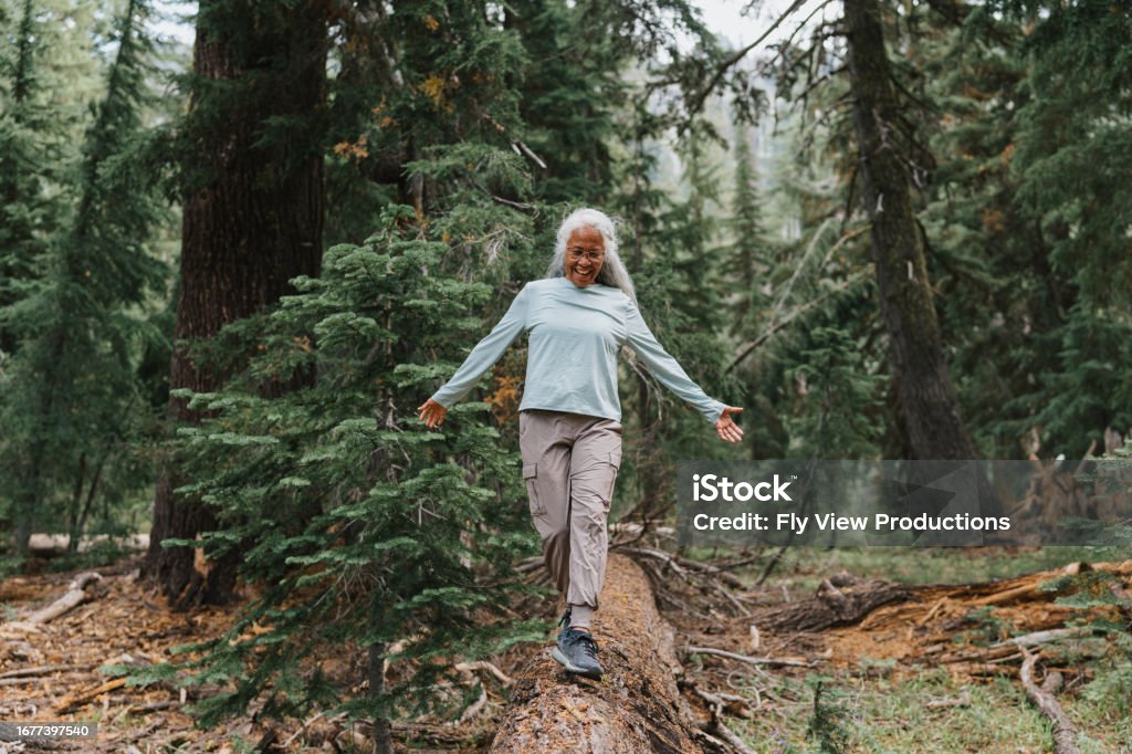 Active senior woman balances on fallen tree Active and healthy senior woman of Pacific Islander descent smiles while walking along the tree trunk of a fallen evergreen tree during a hike in the Pacific Northwest. Balance Stock Photo
