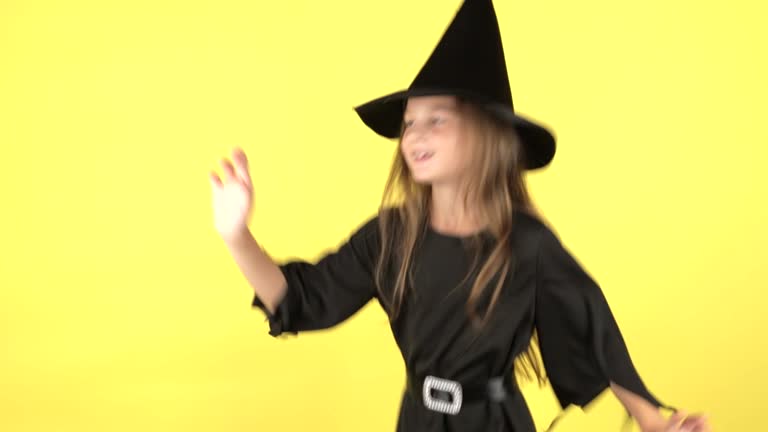 Happy Halloween concept. Portrait of little girl in witch costume raises hands scares and looking at camera, posing isolated on yellow color background wall in studio.