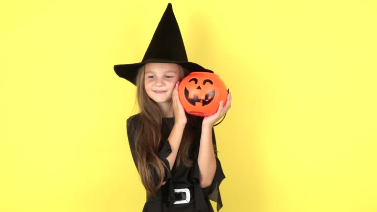 The concept of the holiday Halloween. Portrait smiling little kid girl in black hat witch holding orange pumpkin on face, posing isolated on yellow background in studio.