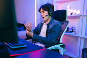 Young Asian esports player man suffering pain in hands and fingers while playing a video game with stress in a bedroom with red and blue neon lights. Concept of pain from gaming addiction and esports.